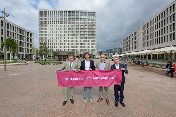 Vier Männer in Anzug halten ein heidelbeerfarbenes Banner mit der Aufschrift "Willkommen am Europaplatz"