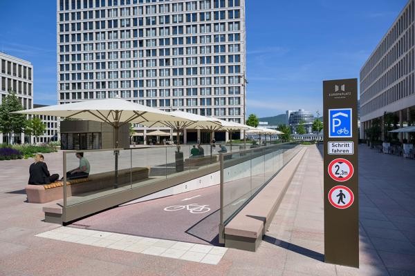 Vom Europaplatz führt eine Rampe bequem ins neue Fahrradparkhaus, das direkt am Heidelberger Hauptbahnhof liegt (im Hintergrund). Das Parken in den ersten 24 Stunden ist kostenfrei. (Foto: Rothe) Eine Rampe führt, auf einem offenen Platz, ins Innere des Fahrradeparkhauses, dahinter ein großes gläsernes Gebäude.