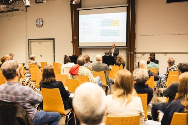 Eine Gruppe von Menschen sitzt in Reihen und betrachtet vor sich eine Präsentation die von einem Mann gegeben wird