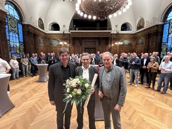 Oberbürgermeister Eckart Würzner verabschiedet Gerald Dietz, den langjährigen Leiter der Geschäfts-stelle Bahnstadt, in den Ruhestand. Drei Personen im Vordergrund, große Personengruppe im Hintergrund