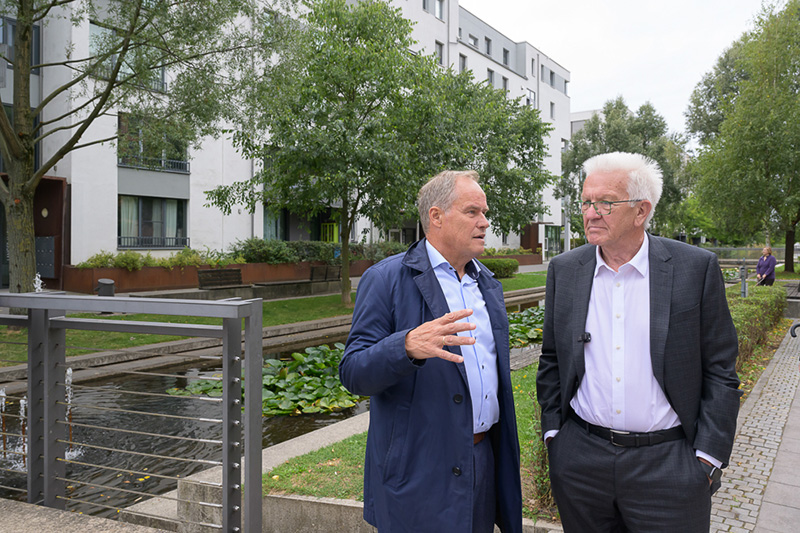 Oberbürgermeister Eckart Würzner (links) mit Ministerpräsident Winfried Kretschmann vor den Wasserbecken im Langer Anger, die zu einem Identifikationsmerkmal des Stadtteils geworden sind Zwei  Männer reden vor dem Hintergrund eines modernen Gebäudes und eines Parks miteinander.