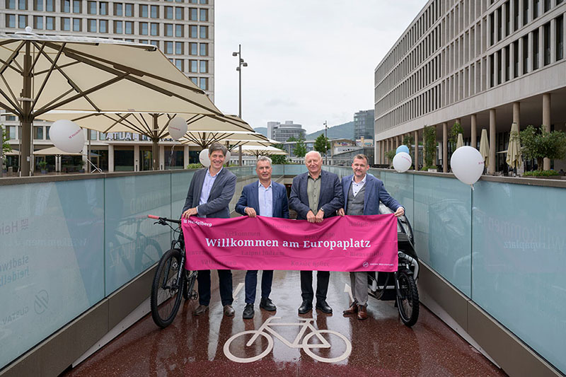 Willkommen im neuen Fahrradparkhaus (von links): Erster Bürgermeister Jürgen Odszuck, Kai Dreesbeimdiek, Geschäftsführer der Gustav Zech Stiftung Management GmbH, Michael Jäger, Geschäftsführer der Stadtwerke Heidelberg Garagen GmbH, und Mobilitätsbürgermeister Raoul Schmidt-Lamontain begrüßen die Radfahrenden an der Fahrradrampe am Europaplatz. (Foto: Rothe) Vier Herren in Jackett posieren, im Eingang eines Fahrradparkhauses, mit einem pinken Banner mit der Aufschrift "Willkommen am Europaplatz."